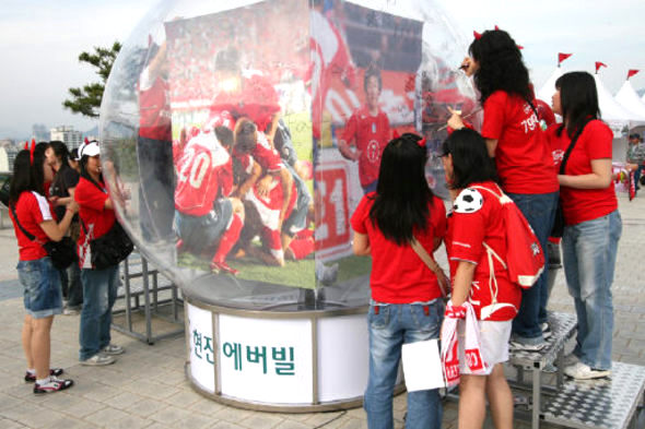 부산 정관 새도시에서 아파트를 분양할 예정인 ㈜현진이 부산 아시아드 주경기장 앞에 축구공 모양의 투명 유리를 설치하고 분양을 홍보하고 있다. 응원용 티셔츠를 선물로 받은 행인들이 유리에 한국대표팀의 선전을 기원하는 사인을 하고 있다. 
현진 제공