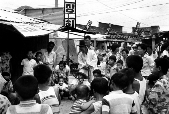 1970년 경북 영주의 한 시장통 사진이다. 식료품 등 각종 잡다한 가게가 늘어서 있는 큰길에서 남녀노소가 무언가를 쳐다보고 있다. 나는 흰 한복을 입은 노인의 모습에 마음이 끌려 자연스럽게 셔터를 눌렀다. 오랜 시간이 흘러 사진을 다시 보니 이들이 무엇을 보고 있었는지가 궁금하다. 유감스럽게도 이 장면은 한장밖에 찍혀 있지 않았다. 보도사진의 기본은 ‘언제 어디서 누가 무엇을 했는지’이지만, 그때의 나는 피사체로부터 받은 감동을 정신없이 사진에 담기에만 열중했다. 한국인의 모습을 찍기 시작한 지 벌써 반세기가 되려 하고 있다. 최근 사진을 정리하고 문서로 기록을 남기는 것의 중요성을 느끼고 있다. 후지모토 다쿠미/사진가