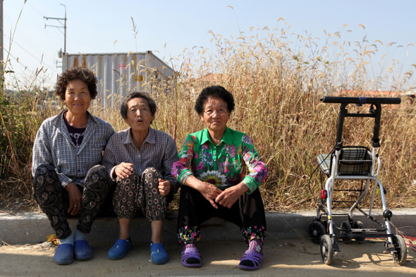 평택시 고덕면 율포리의 한 마을이 ‘고덕 국제 신도시 건설지구’에 포함됐다. 마을 주민은 떠나야 했다. 선택권은 없었다. 이들을 위해 나라에선 인근 지역에 ‘율곡 타운’을 조성해 이주를 도왔다. 불편하고 오래된 것을 버리고 새것을 얻었지만, 그사이 잊혀진 것들도 많다. 구불구불한 마을 길, 늘 그 자리에 있던 수목(樹木), 텃밭, 나고 자란 고향의 추억, 그리고 절반의 이웃들…. 율포리 마을 주민의 입주를 위해 건축되고 있는 현장을 먼저 정착한 마을 어르신들이 바라보고 있다. 2014년 10월.  
백홍기/사진가 