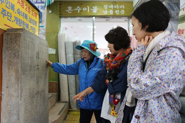‘이야기 할매’ 문영선(왼쪽)씨가 탐방객들에게 부산 국제시장의 한 이불가게 앞에 서 있는 상가 ‘준공 기념비’(1970년)에 대해 설명하고 있다.