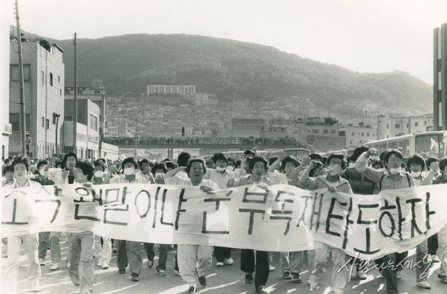 1987 6월10일 (박종철 고문치사사건) “고문살인 은폐조작 웬 말이냐 군부독재 타도하자”라는 펼침막을 들고 시위대 앞에서서 구호를 외치는 노무현 변호사와 부산시민들.  노무현재단(노무현 사료관) 누리집